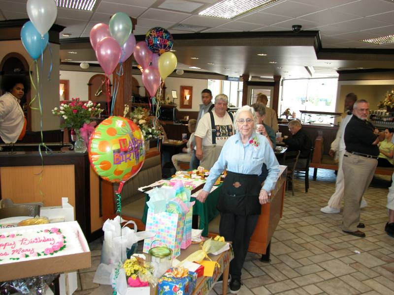 Mary LeForge surrounded by two cakes, banners, balloons and a table full of gifts and cards from her friends and customers